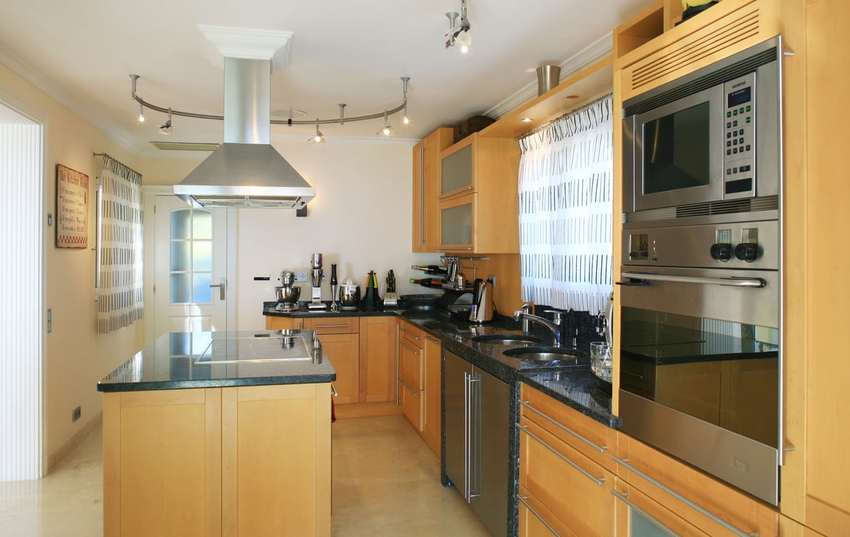 Cuisine moderne avec armoires en bois clair, comptoirs en granit foncé et îlot central intégrant une table de cuisson. Appareils en acier inoxydable : four, micro-ondes et hotte.