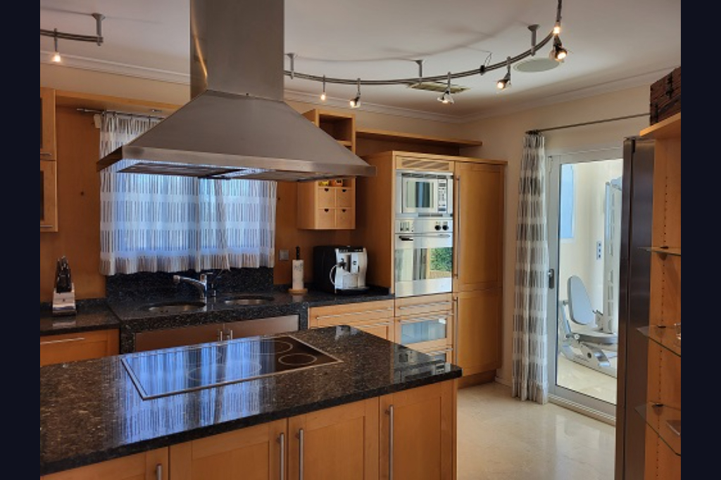 Cuisine moderne avec des armoires en bois clair, des plans de travail en granit foncé, un îlot avec plaque de cuisson intégrée et des appareils en acier inoxydable. Accès à une salle de sport.