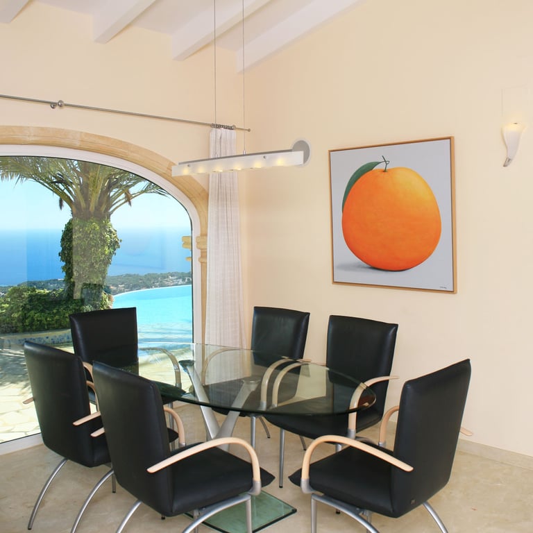 Espace repas avec table en verre et chaises modernes, agrémenté d'une grande fenêtre arquée offrant des vues panoramiques sur la mer et la côte. Une œuvre d'art d'orange décore le mur.