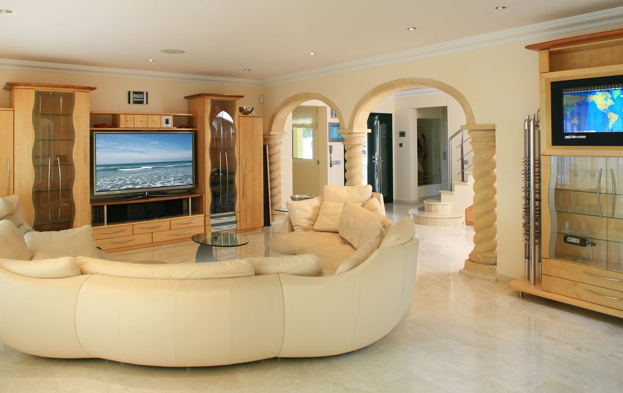 Grand salon avec canapé incurvé crème, sols en marbre clair et meubles TV en bois clair sur mesure. Des arches en pierre avec colonnes mènent à l'entrée et à l'escalier.