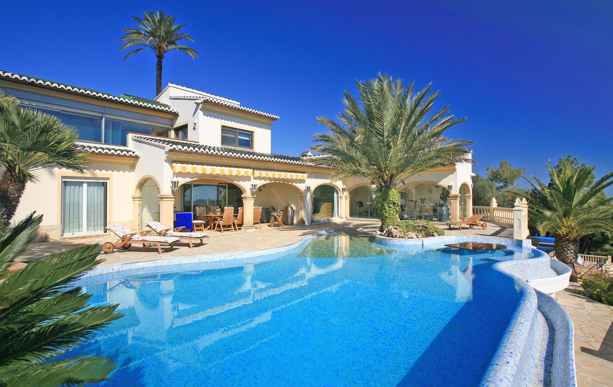 Grande piscine en mosaïque bleue avec entrée plage, îlot de palmiers et table aquatique. La villa propose une naya couverte et une salle de sport vitrée.