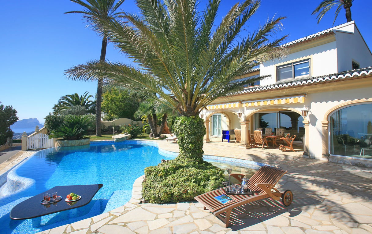 La piscine au design unique intègre une table immergée pour les rafraîchissements, bordée d'une terrasse en pierre et de palmiers, avec vue mer lointaine.