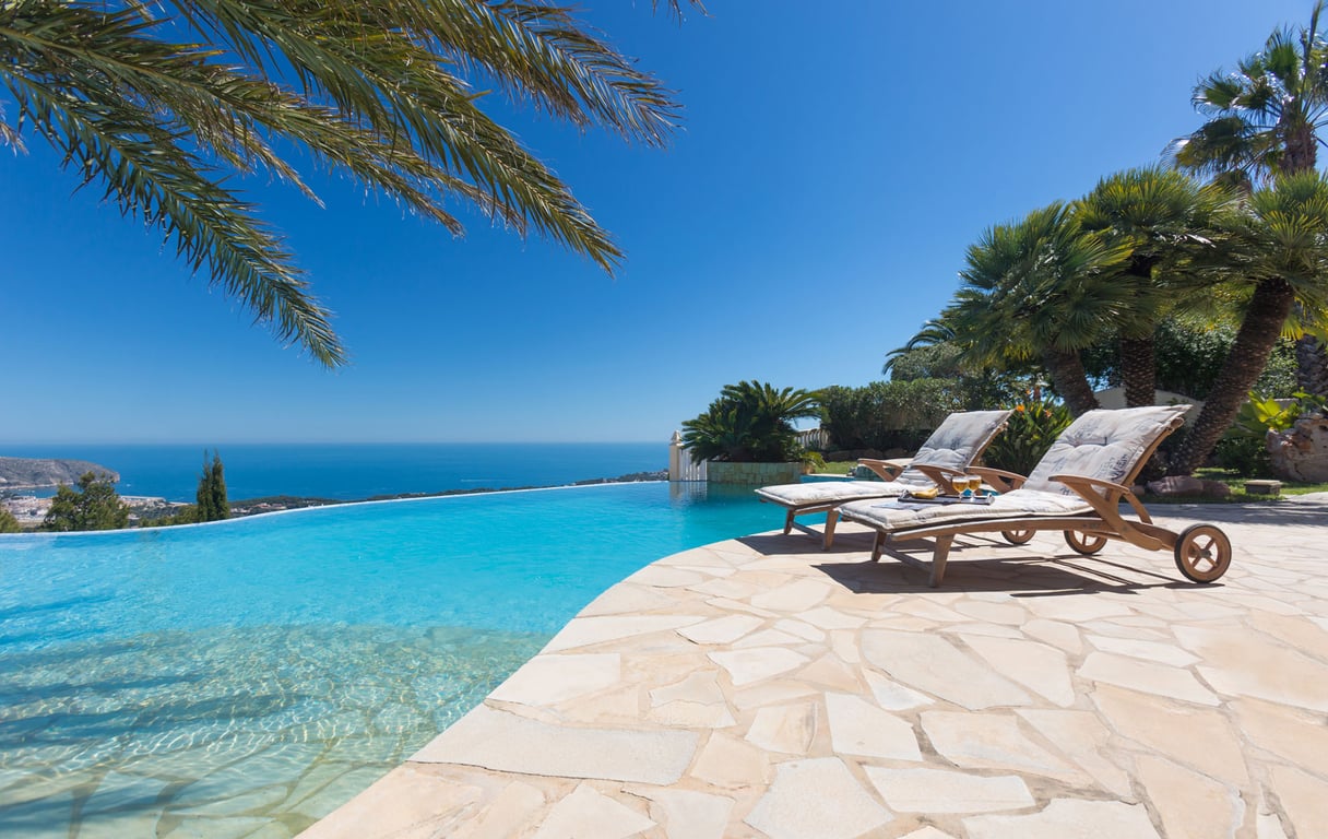La terrasse de la piscine à débordement présente deux chaises longues en bois sur un patio en dalles, avec une vue imprenable sur la mer Méditerranée et la côte.