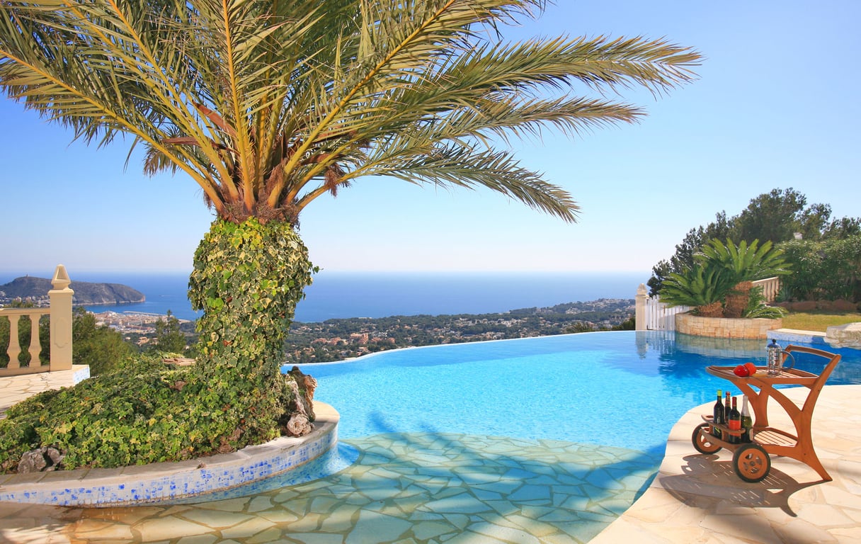 Piscine à débordement avec vue panoramique sur la mer et la côte, agrémentée d'un palmier recouvert de lierre et d'un chariot de bar en bois sur la terrasse.