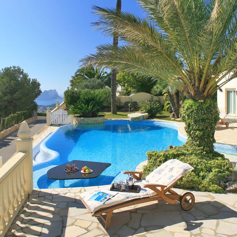 Piscine incurvée avec une table noire immergée et des rafraîchissements, jouxtant une terrasse pavée de pierre avec transat. Vues lointaines sur la mer et une formation rocheuse distincte.