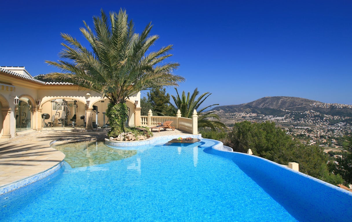 Piscine incurvée en mosaïque bleue avec îlot de palmiers et table immergée. Salle de sport vitrée et terrasse offrant des vues panoramiques sur la montagne.
