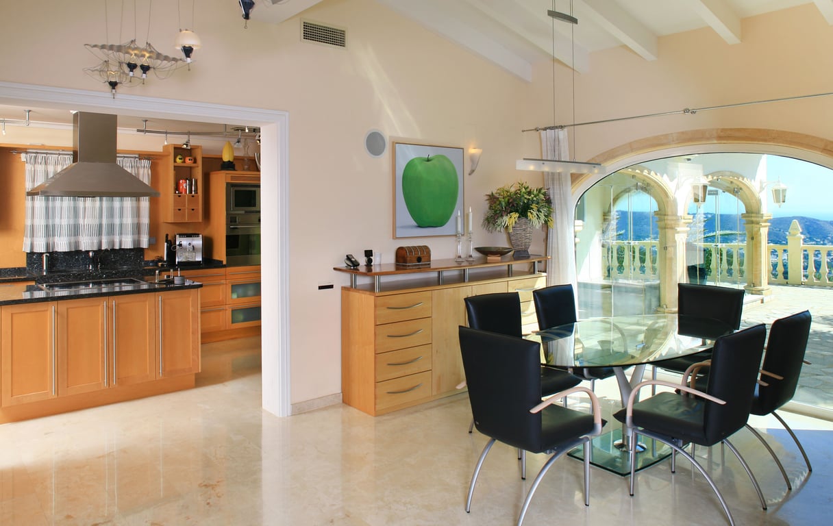 Salle à manger lumineuse avec table en verre et chaises noires, sols en marbre poli. De grandes arches vitrées offrent des vues panoramiques sur la mer et les montagnes, attenante à une cuisine moderne.
