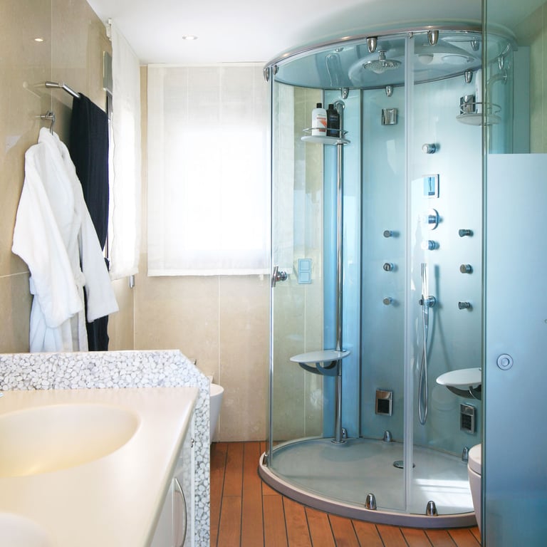 Salle de bain avec cabine de douche en verre à jets multiples, un plan de travail de lavabo texturé de galets et un sol en bois chaleureux. Lumière naturelle par la fenêtre.