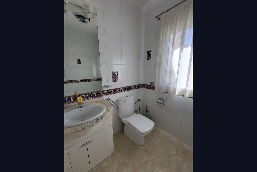 Salle de bain avec murs carrelés blancs, frise décorative et carreaux à motif coq. Elle dispose d'un lavabo sur meuble à plateau en marbre clair, de toilettes et d'une fenêtre.