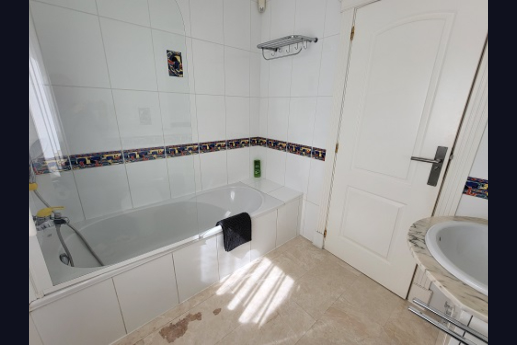 Salle de bain fonctionnelle avec baignoire blanche, pare-douche en verre et murs carrelés de blanc avec frise décorative. Dispose d'un lavabo en marbre et lumière naturelle.
