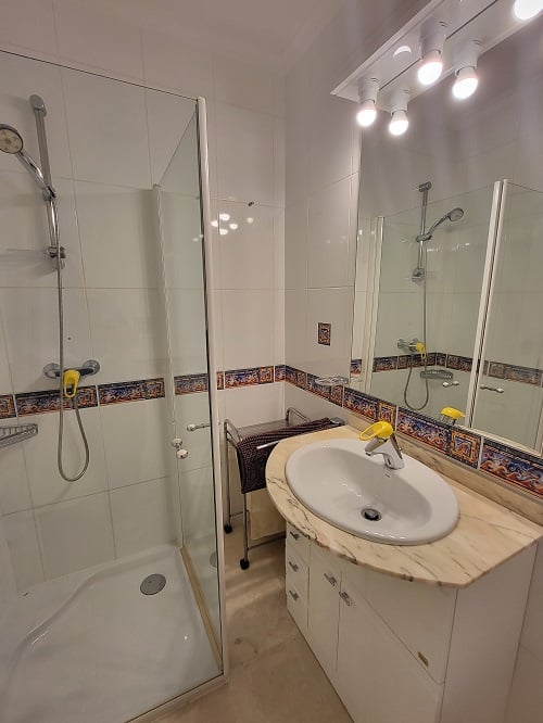 Salle de bain fonctionnelle avec cabine de douche vitrée, meuble-lavabo à plateau en marbre clair et miroir éclairé. Carrelage blanc avec frise méditerranéenne décorative.