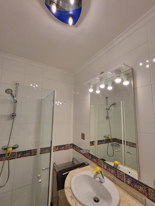 Salle de bain fonctionnelle avec carrelage blanc et frise décorative, douche vitrée et lavabo sur meuble effet marbre. Miroir éclairé et plafonnier bleu et argent.