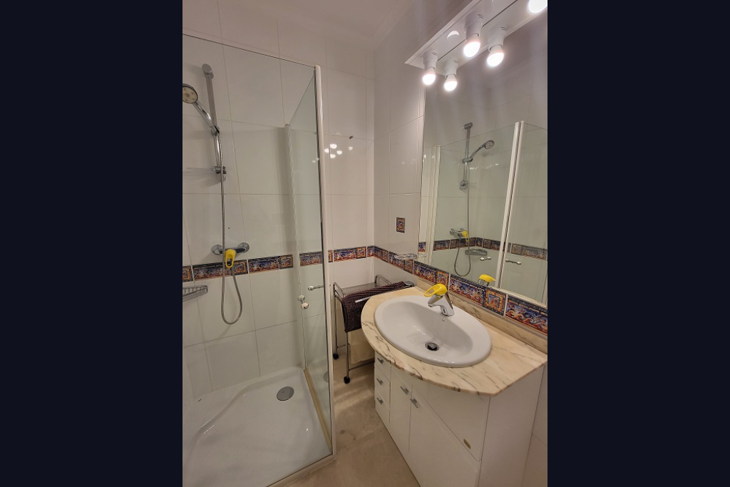 Salle de bain fonctionnelle avec douche vitrée, lavabo sur plan de travail en marbre clair et meuble blanc. Les murs carrelés blancs sont rehaussés d'une frise décorative méditerranéenne.