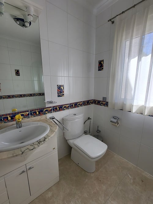 Salle de bain lumineuse avec carrelage mural blanc, frise décorative colorée et accents. Comprend une vasque sur meuble en marbre, des toilettes modernes et une fenêtre avec voilage.