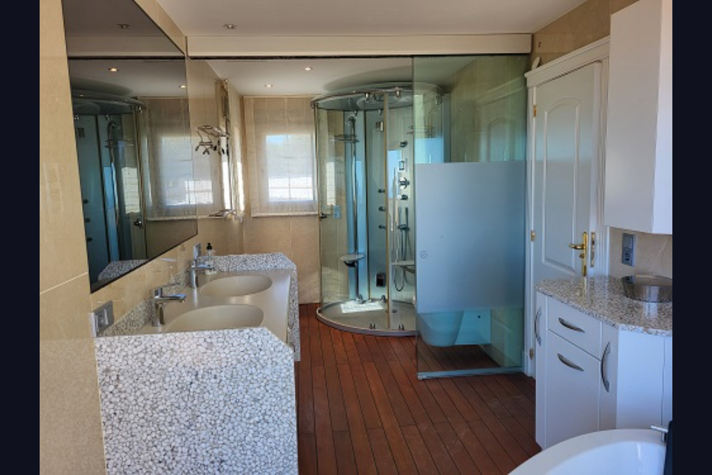 Salle de bain spacieuse avec double vasque au design galet, douche hydromassante circulaire et parquet. Grand miroir et fenêtres avec stores visibles.