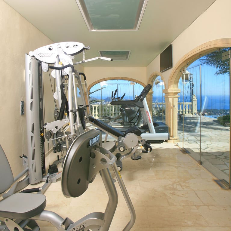 Salle de sport privée avec équipements Hoist et Life Fitness, sol en marbre et baies vitrées arquées sur la mer et la piscine. Des puits de lumière complètent l'éclairage naturel.