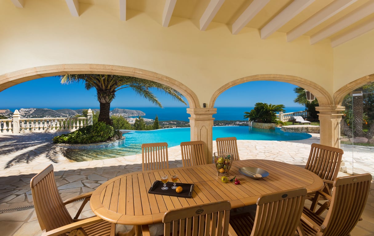 Vaste naya couverte avec table et chaises en bois, offrant une vue panoramique sur la mer Méditerranée et la côte, encadrée par des arches en pierre et une piscine à débordement.