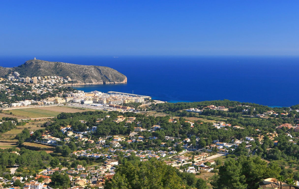 Vue panoramique depuis une colline, présentant une ville côtière, un port de plaisance, une montagne proéminente et la mer Méditerranée sous un ciel clair.