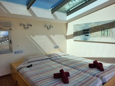 Chambre lumineuse avec deux lits simples, parures de lit rayées et serviettes rouges. Un puits de lumière zénithal éclaire la pièce, agrémenté d'un tableau abstrait et d'une fenêtre.