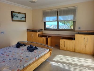 Chambre lumineuse avec lit en bois clair, bureau intégré et rangements. Les fenêtres offrent des vues panoramiques sur la côte, inondant la pièce de lumière naturelle.