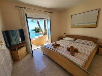 Chambre lumineuse avec lits jumeaux et tête de lit en osier, offrant un accès direct à la piscine et des vues sur la mer. Télévision et œuvre d'art murale.