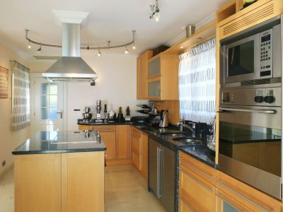 Cuisine moderne avec armoires en bois clair, comptoirs en granit foncé et îlot central intégrant une table de cuisson. Appareils en acier inoxydable : four, micro-ondes et hotte.