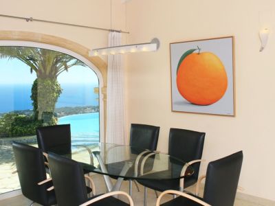 Espace repas avec table en verre et chaises modernes, agrémenté d'une grande fenêtre arquée offrant des vues panoramiques sur la mer et la côte. Une œuvre d'art d'orange décore le mur.