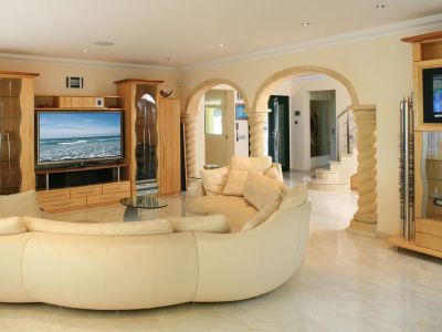 Grand salon avec canapé incurvé crème, sols en marbre clair et meubles TV en bois clair sur mesure. Des arches en pierre avec colonnes mènent à l'entrée et à l'escalier.