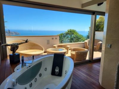Jacuzzi intérieur sur parquet en bois s'ouvrant sur une terrasse privée avec mobilier en rotin, offrant une vue panoramique sur la mer Méditerranée.