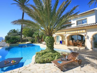 La piscine au design unique intègre une table immergée pour les rafraîchissements, bordée d'une terrasse en pierre et de palmiers, avec vue mer lointaine.