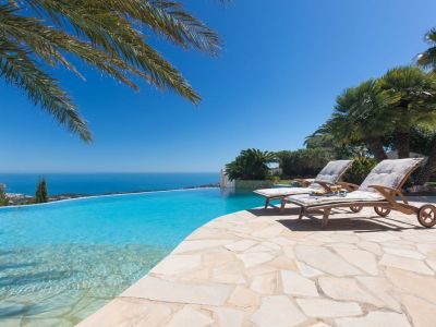 La terrasse de la piscine à débordement présente deux chaises longues en bois sur un patio en dalles, avec une vue imprenable sur la mer Méditerranée et la côte.