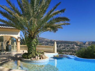 Piscine à débordement avec île de palmiers et table intégrée, offrant des vues panoramiques sur la côte et les montagnes sous un ciel clair.