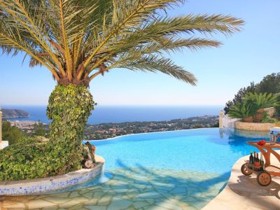 Piscine à débordement avec vue panoramique sur la mer et la côte, agrémentée d'un palmier recouvert de lierre et d'un chariot de bar en bois sur la terrasse.
