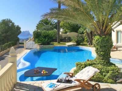 Piscine incurvée avec une table noire immergée et des rafraîchissements, jouxtant une terrasse pavée de pierre avec transat. Vues lointaines sur la mer et une formation rocheuse distincte.