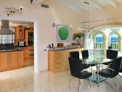 Salle à manger lumineuse avec table en verre et chaises noires, sols en marbre poli. De grandes arches vitrées offrent des vues panoramiques sur la mer et les montagnes, attenante à une cuisine moderne.