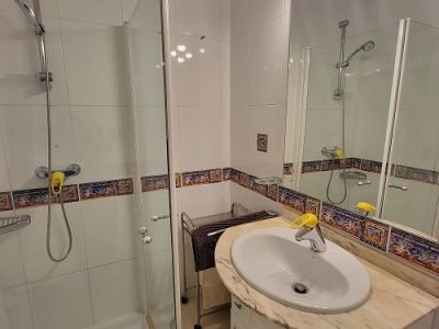 Salle de bain fonctionnelle avec cabine de douche vitrée, meuble-lavabo à plateau en marbre clair et miroir éclairé. Carrelage blanc avec frise méditerranéenne décorative.