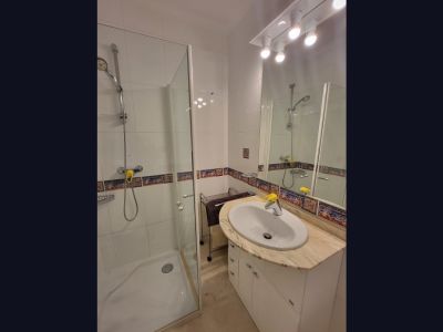 Salle de bain fonctionnelle avec douche vitrée, lavabo sur plan de travail en marbre clair et meuble blanc. Les murs carrelés blancs sont rehaussés d'une frise décorative méditerranéenne.