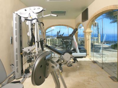Salle de sport privée avec équipements Hoist et Life Fitness, sol en marbre et baies vitrées arquées sur la mer et la piscine. Des puits de lumière complètent l'éclairage naturel.