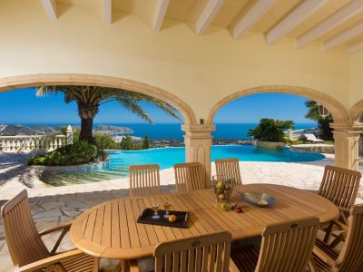 Vaste naya couverte avec table et chaises en bois, offrant une vue panoramique sur la mer Méditerranée et la côte, encadrée par des arches en pierre et une piscine à débordement.