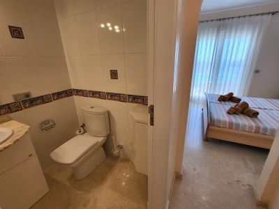 Vue depuis une salle de bain carrelée avec toilettes et lavabo, donnant sur une chambre lumineuse avec lits jumeaux, linge de lit rayé et grande fenêtre voilée.