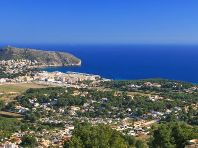 Vue panoramique depuis une colline, présentant une ville côtière, un port de plaisance, une montagne proéminente et la mer Méditerranée sous un ciel clair.