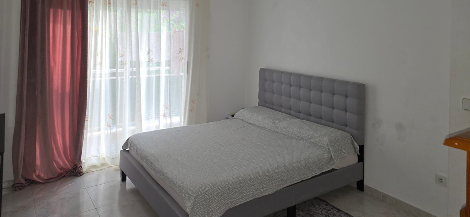 Chambre à coucher avec un lit gris capitonné et une literie claire à motifs. La lumière naturelle pénètre par la grande fenêtre, habillée de voilages et de rideaux bordeaux, sur un sol carrelé.