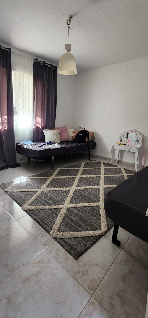Chambre avec carrelage clair, un tapis géométrique gris et beige, et un lit simple près de la fenêtre avec rideaux sombres. Une petite coiffeuse blanche est présente.