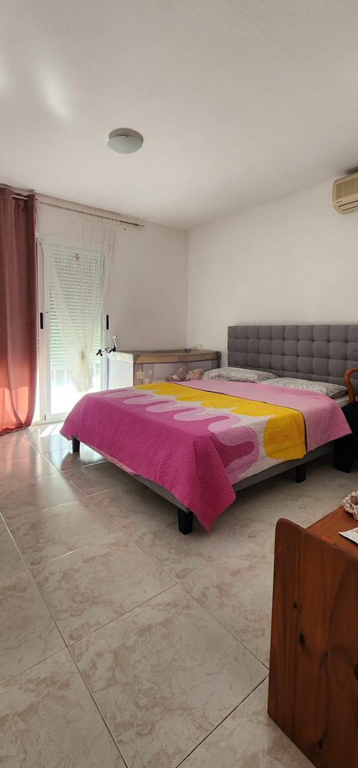 Chambre lumineuse avec lit double, tête de lit capitonnée grise et sol carrelé. Accès direct à l'extérieur et climatisation murale.