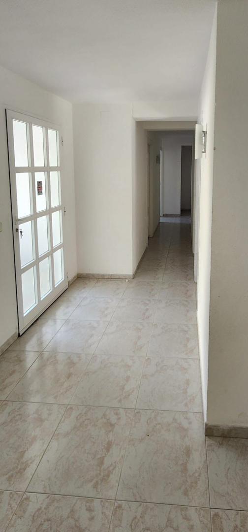 Couloir intérieur avec un sol carrelé clair et des murs blancs. Une porte blanche à panneaux de verre dépoli se trouve à gauche, éclairée par une applique murale le long du couloir.