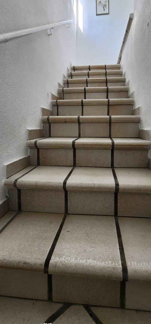 Escalier intérieur avec des marches carrelées de couleur claire et des joints foncés, éclairé par la lumière naturelle d'une fenêtre supérieure. Mains courantes blanches des deux côtés.