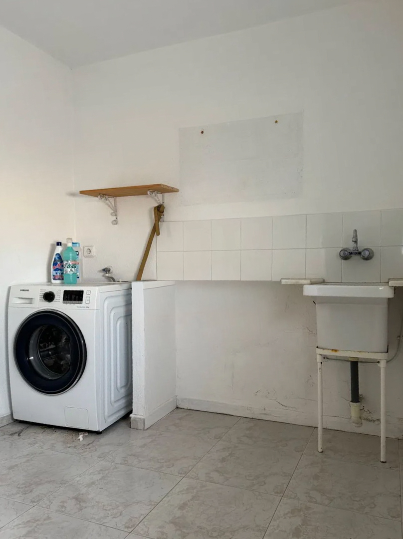 Espace buanderie fonctionnel avec machine à laver moderne, évier utilitaire simple et crédence carrelée blanche. Le sol est en carrelage clair, complété par une étagère en bois.