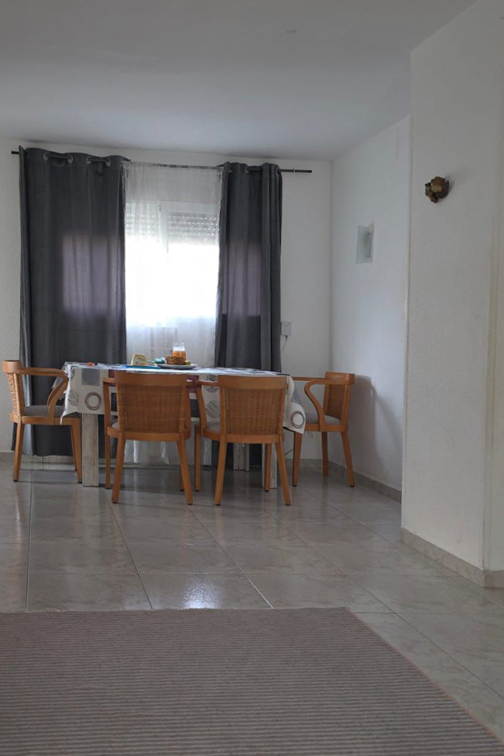 Espace salle à manger lumineux avec table en bois et chaises tressées, situé près d'une fenêtre aux rideaux gris. Le sol est carrelé de couleur claire, avec un tapis rayé au premier plan.