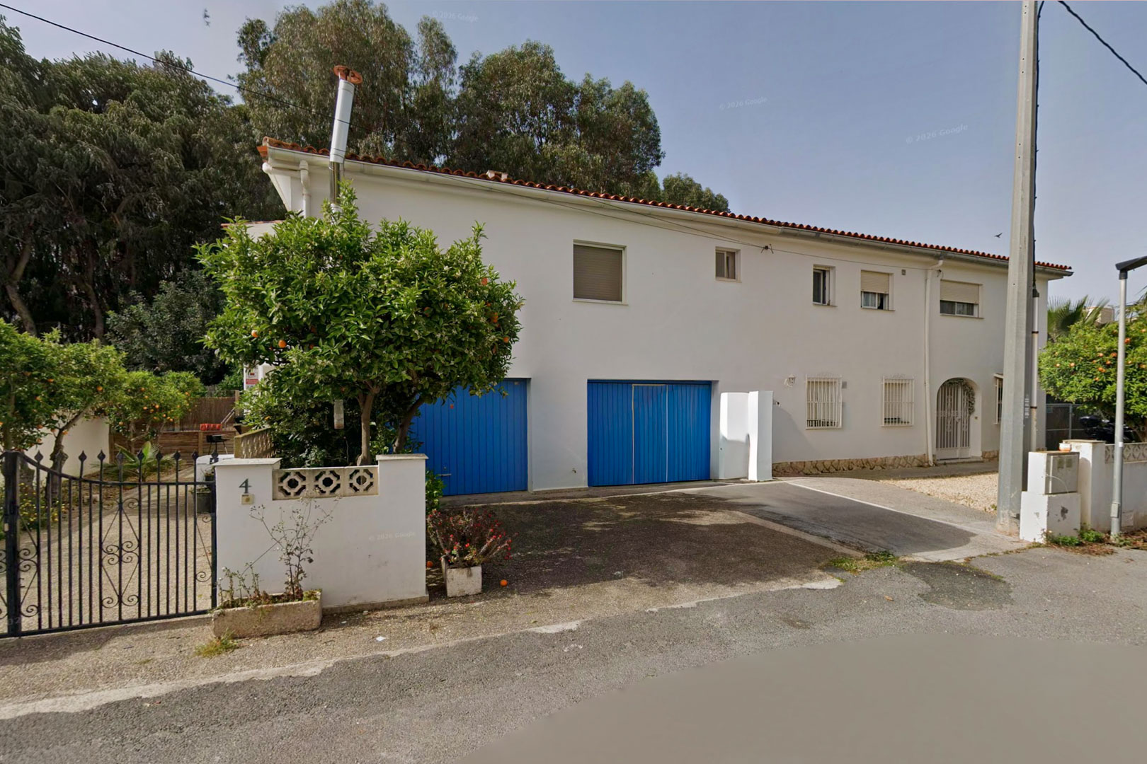 Façade avant de la villa jumelée, présentant une façade blanche, deux grandes portes de garage bleues et un oranger près du portail en fer forgé.