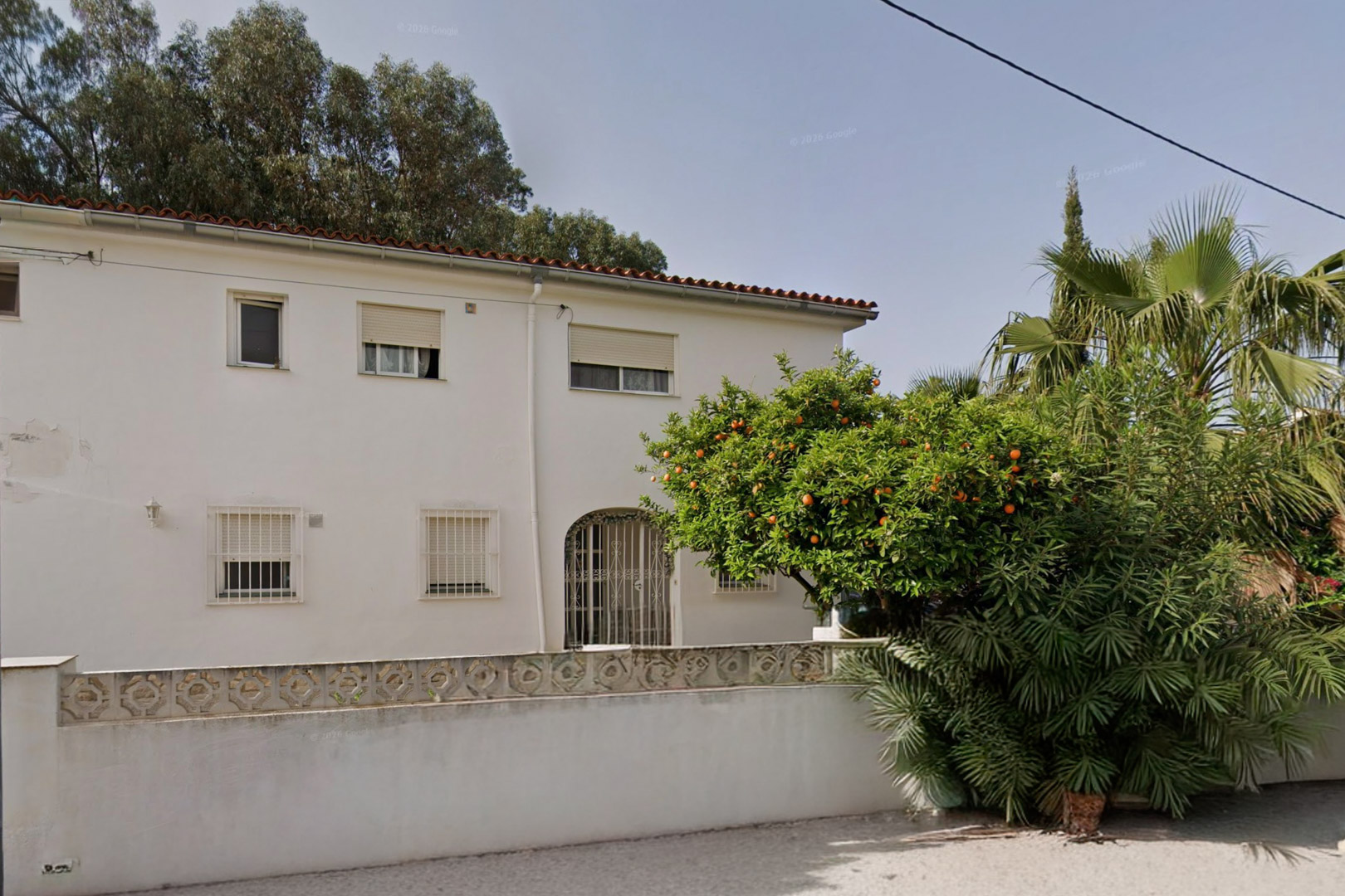 Façade blanche de la villa avec toit en tuiles rouges, fenêtres à barreaux et entrée arquée. Un oranger fructueux et une végétation luxuriante embellissent le jardin avant.